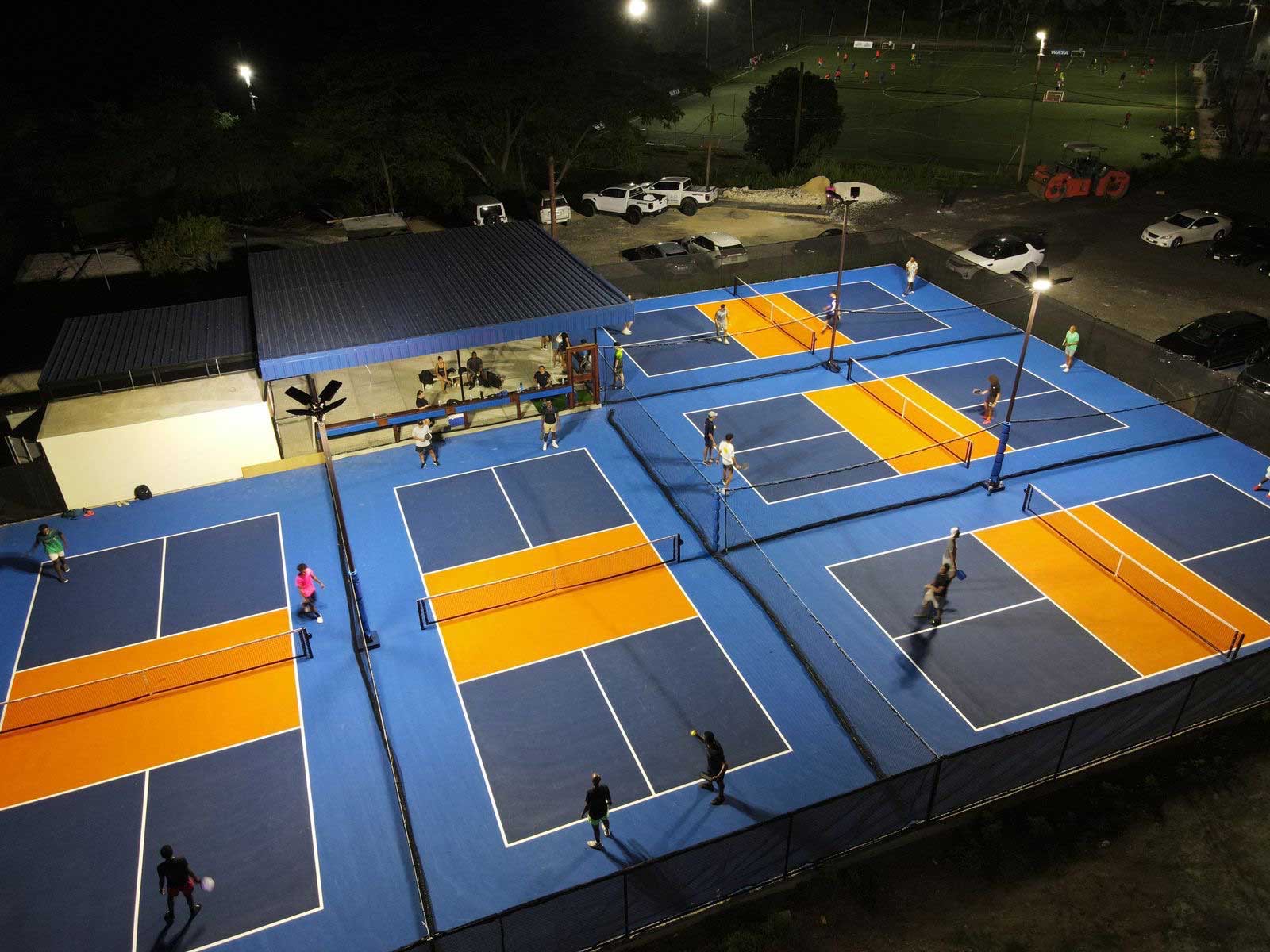 Lit blue and orange pickleball courts at night, full of players